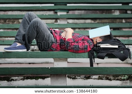 an asian boy sleeping on a green concrete bench with blue book covering his face