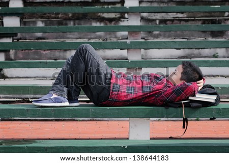 an asian boy sleeping on a green concrete bench with backpack under his head
