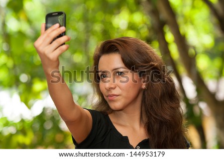 Young attractive Hispanic woman portrait outside green behind taking her own picture