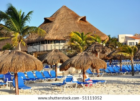 In the Mexican style. A resort building with a grassy roof on seacoast. Dark blue plank beds, sand, the umbrellas protecting from the sun.