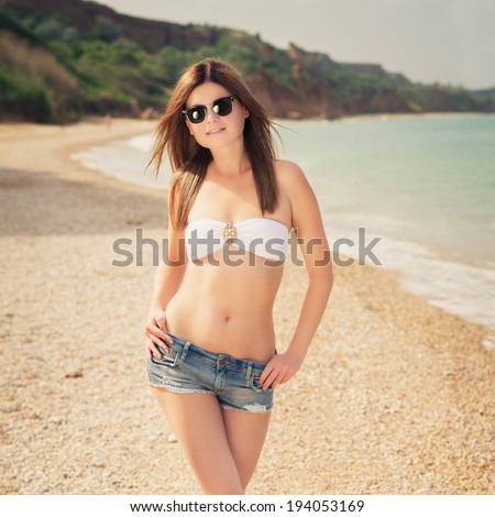 Outdoor fashion warm colors portrait of young sensual happy woman in jeans shorts, bikini and sunglasses. model posing. Photo in style instagram filters