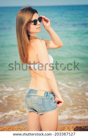 Outdoor fashion warm colors portrait of young sensual happy woman in jeans shorts, bikini and sunglasses. model posing