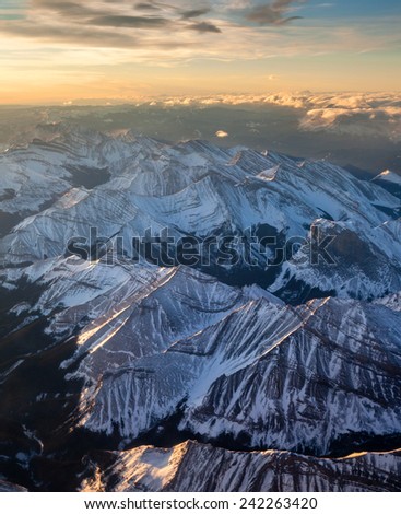 An aerial view of a golden sunrise at the break of dawn over the Canadian Rockies, with snow-capped mountains and clouds on mountain tops.