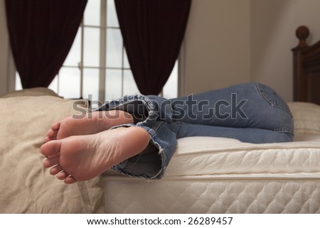 A woman in blue jeans relaxing on the bed during the day with feet hanging off.