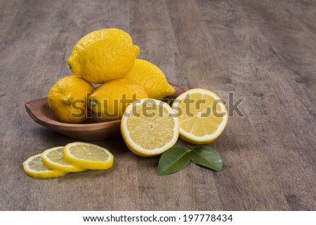 A sliced lemon and a lemon cut in a half in front of a pot of lemons over a wooden surface.