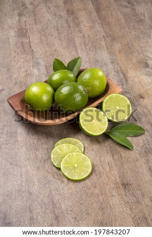 Some entire green lemons in a pot and a lemon cut in a half with leaves and three slices of a lemon over a wooden table.