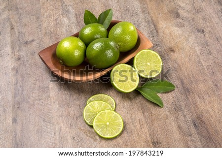 Some entire green lemons in a pot and a lemon cut in a half with leaves and three slices of a lemon over a wooden table.