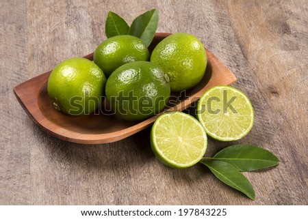 Some entire green lemons in a pot and a lemon cut in a half with leaves over a wooden table.
