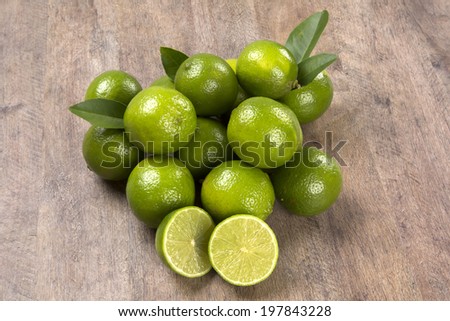 Some entire green lemons with leaves and a lemon cut in a half on a wooden table.