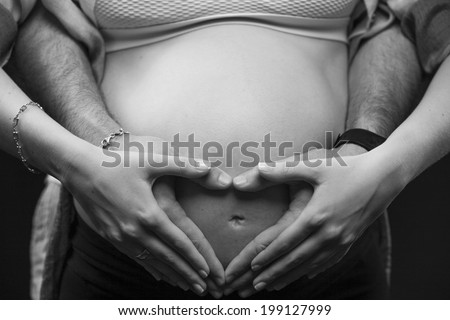 Heart shaped hands of a pregnant woman with her husband in black and white on a black background.