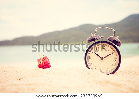 Concept smile alarm clock and small red Christmas gift box on beautiful sand beach. Vintage filter.