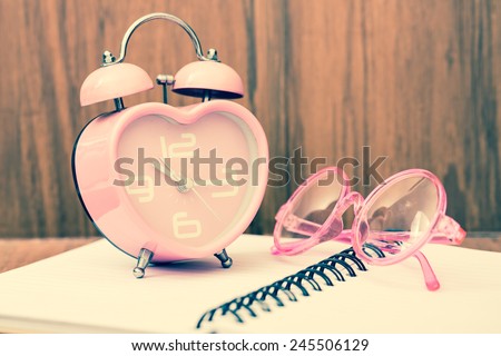 Vintage heart shape alarm clock with chic eye glasses on open notebook. With wood background. Retro filter.