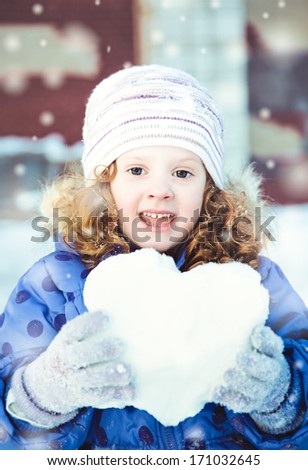 Beautiful little girl happily holding a snowball in the shape of heart  in winter park on bokeh background with snowflakes.