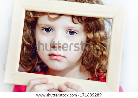Closeup portrait of a beautiful little curly girl with big gray eyes,  holding empty wooden frame.