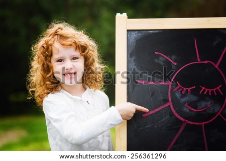 Happy girl points a finger at the picture with an happy sun, painted in red chalk on a blackboard. Educational concept.
