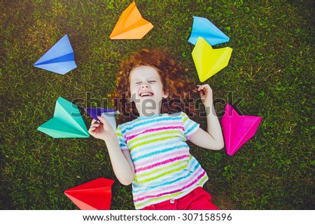 Laughing girl throwing paper airplane in green grass at summer park.