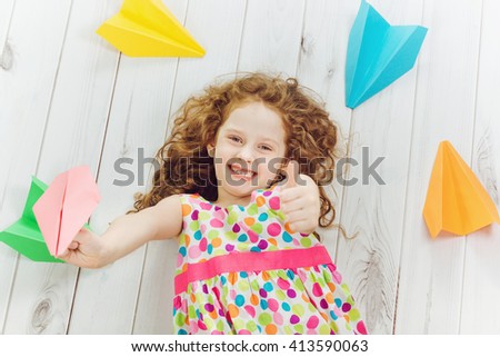 Happy laughing child throwing paper airplane indoors and  showing thumbs up.\
Happy childhood, travel, vacation concept. Top view.