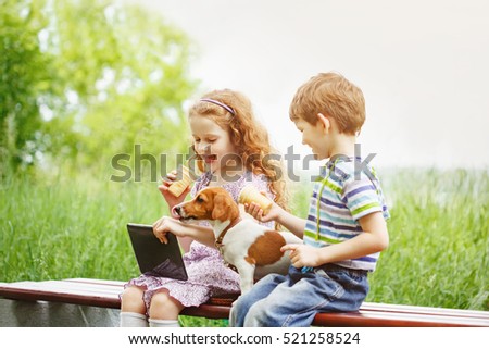 Happy child playing and having fun with her puppy. Girl playing in the tablet PC and eating ice cream in summer park. Leisure, friendship or family concept.