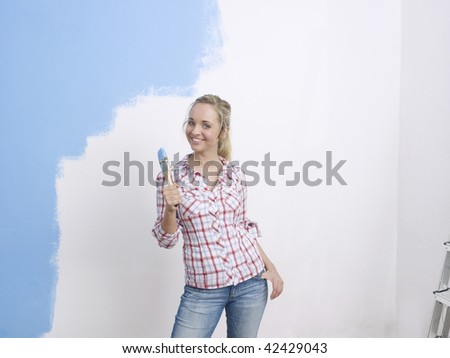 Half body portrait of young woman with paintbrush and partially covered blue wall in background.
