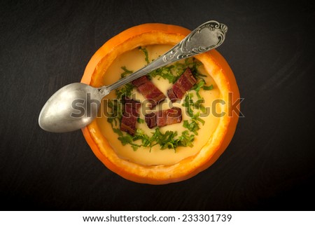 Overhead shot of pumpkin and potatoes soup served inside real pumpkin, decorated with parsley and fried ham.