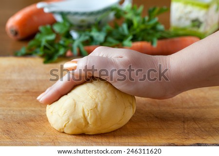 Hand above pasta dough on a cutting board with carrots and parsley in the background.