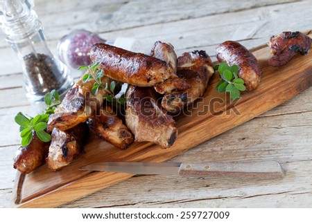 Wooden board with roasted pork ribs and sausages decorated with fresh marjoram, with violet salt and pepper grinder in the background.