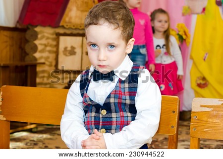 Sad boy in a vest and bow tie seated on a chair at the party in the kindergarten