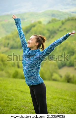 Beautiful young woman outdoor, happy with raised arms enjoying the free time. Spring. Young woman in blue woolen sweater relaxing on nature. Enjoy Nature. Healthy Smiling Girl in Green Grass
