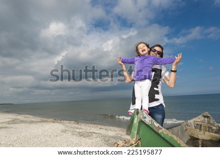 Little girl with mom having fun on the beach in a old boat against blue cloudy sky