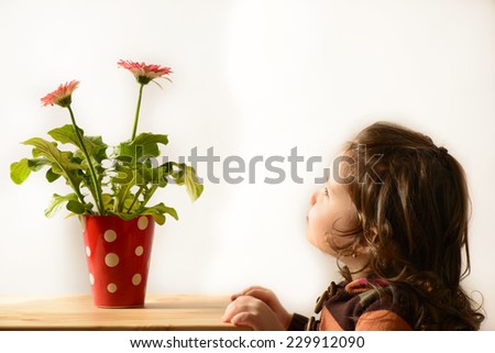 Beautiful little girl sitting next to a table with red vase with flowers on it, studio shot, on white background. Cute little admiring vase with flowers