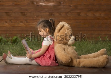 Beautiful little girl sitting back with toy bear outside in backyard playground, reading to her little plush friend
