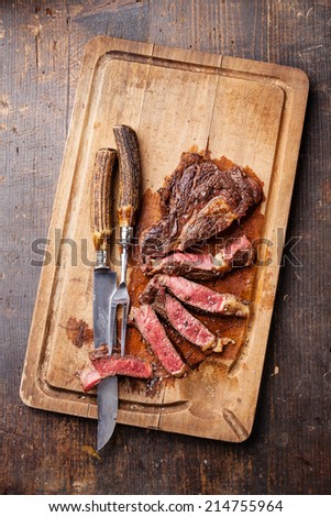 Medium rare grilled Beef steak Ribeye with knife and fork for meat on cutting board on dark wooden background