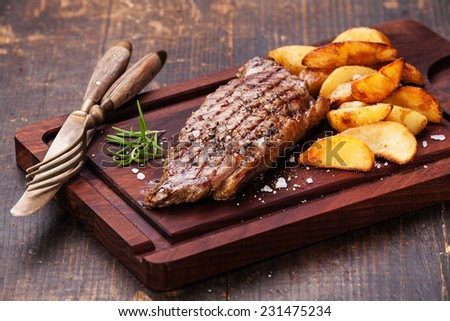 Well done grilled New York steak with roasted potato wedges on cutting board on dark wooden background