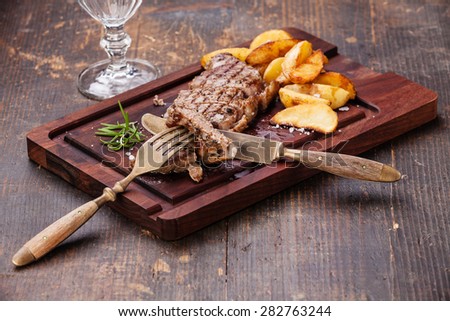Sliced well done grilled New York Steak with roasted potato wedges on cutting board on dark wooden background