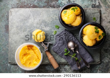 Mango ice cream sorbet with mint leaves in black bowls, Spoon for ice cream and plastic container on stone slate background