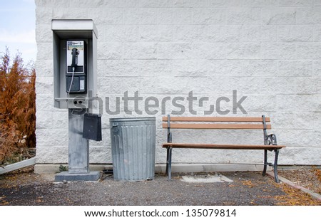An old pay phone next to a metal trashcan and a wooden bench in front of a white painted cinderblock wall.