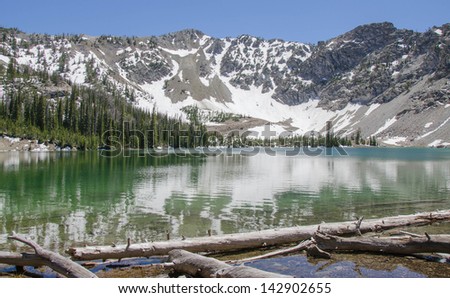Pristine Idaho mountain lake in the Sawtooth National Forest with trees and snow covered peaks in the background reflected upon calm water.