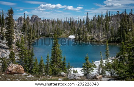Blue lake in Idaho Sawtooth Wilderness surrounded by trees with blue sky and white clouds above.