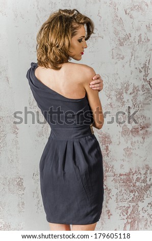 Young beautiful brown-haired woman in a blue dress with bare shoulders and backs against the wall and worth looking dreamily