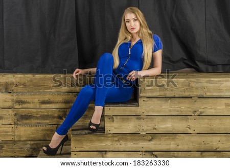 Beautiful long haired blonde in blue overalls and high heels sitting on wooden stairs