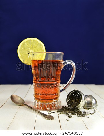 Glass cup of cold tea with lemon slice, old spoon and raw tea leaves in strainer on blue background, vintage still life