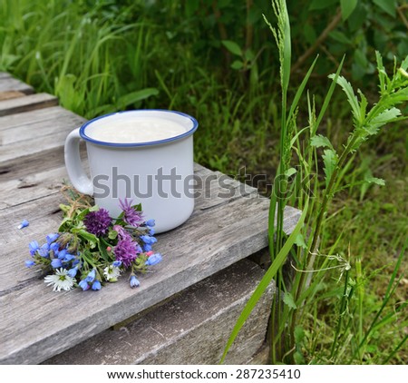 Cup of milk with bunch of wildflowers on old wooden table over grass background, summer rural still life