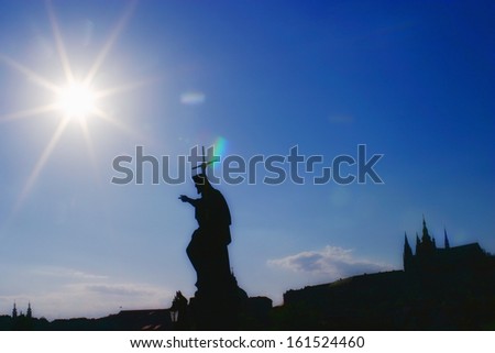 A statue of god holding a cross, with a building in the distance and the sun shining.