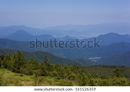 A green mountain side looking into a valley with a mountain range in the background.