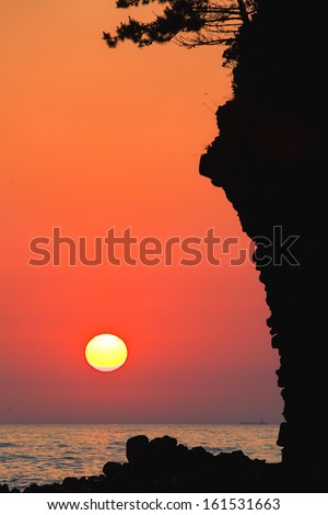 A rocky cliff perched over the ocean with a purple, red and yellow sunset.