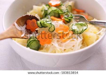 A bowl of vegetables and rice noodles with a serving fork and a wooden spoon.