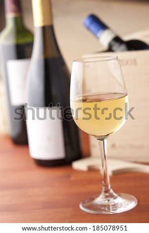 A wine glass filled half way with white wine, three assorted bottles of wine behind it.