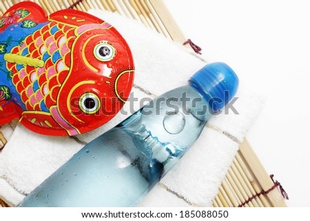 A glass water bottle and fish-shaped metal toy resting on a napkin and a bamboo mat.