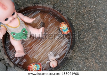 A dolls sitting among other toys in water in a round wooden bucket.