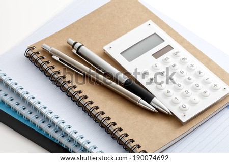 Two pens and a calculator sit on top of three notebooks.